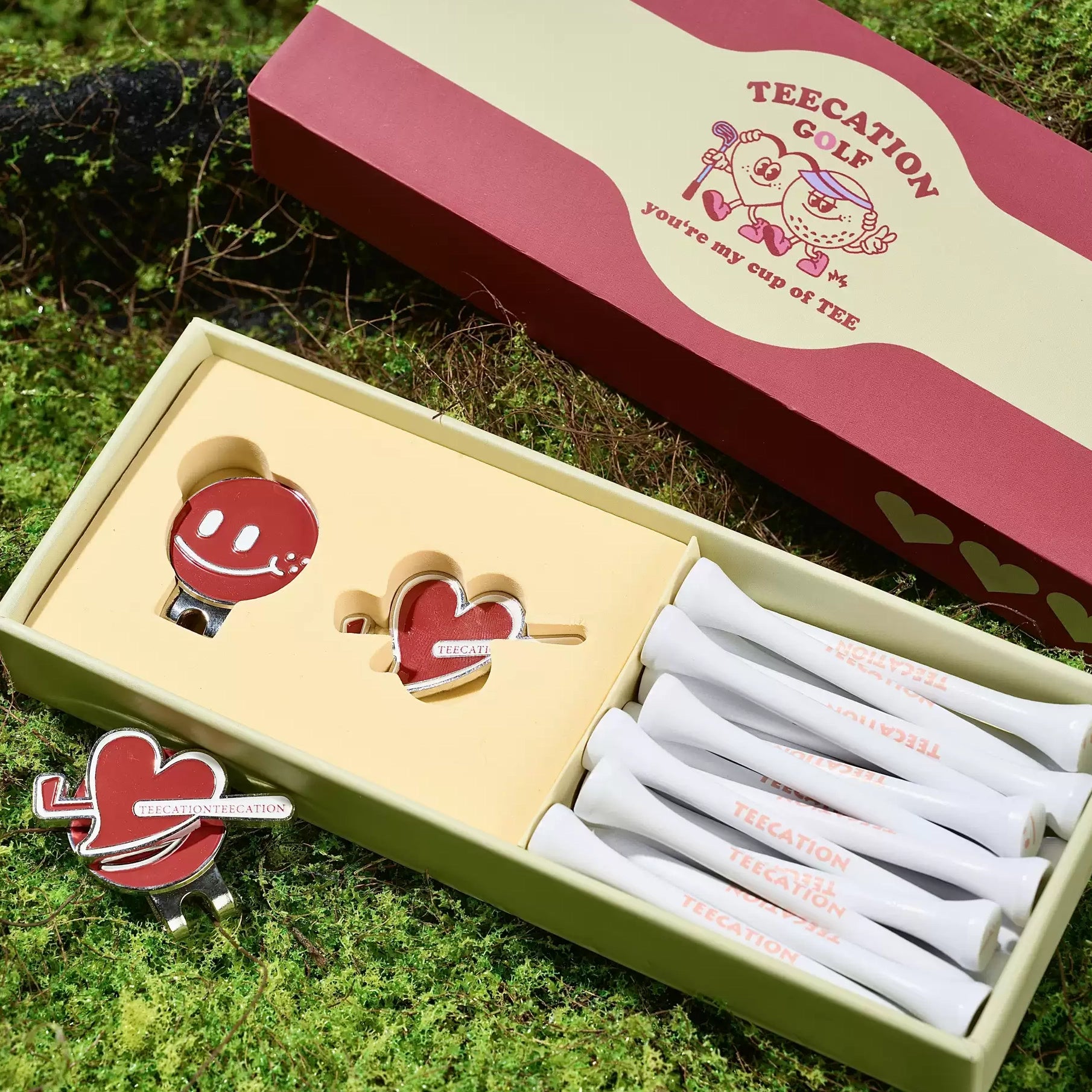 A TEECATION GOLF gift set displayed on green grass, featuring a red and cream branded box, a light green tray holding white golf clubs, and matching red and cream accessories.