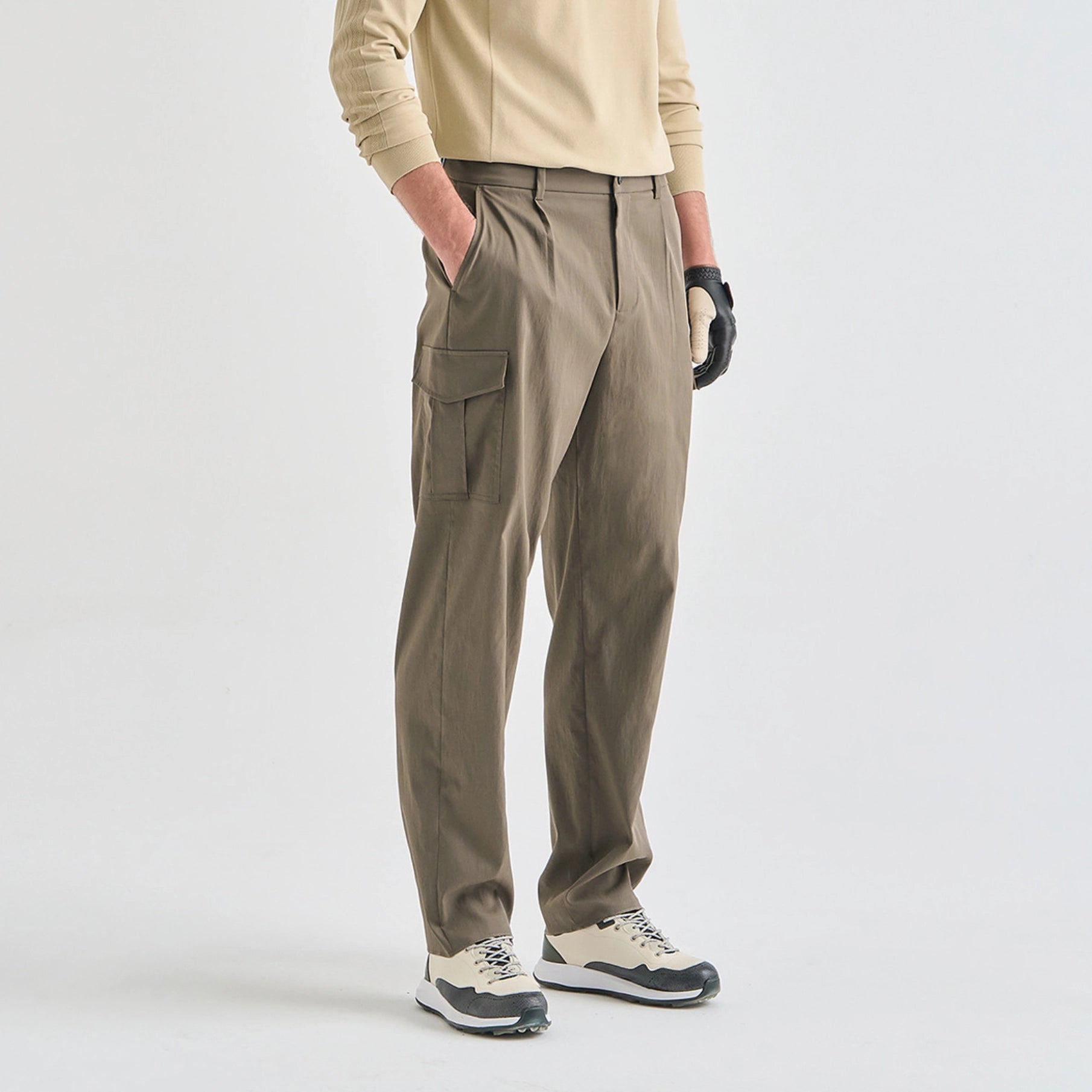 A model wearing khaki cargo-style trousers, a beige long-sleeve top, and golf shoes, standing against a light gray studio background.