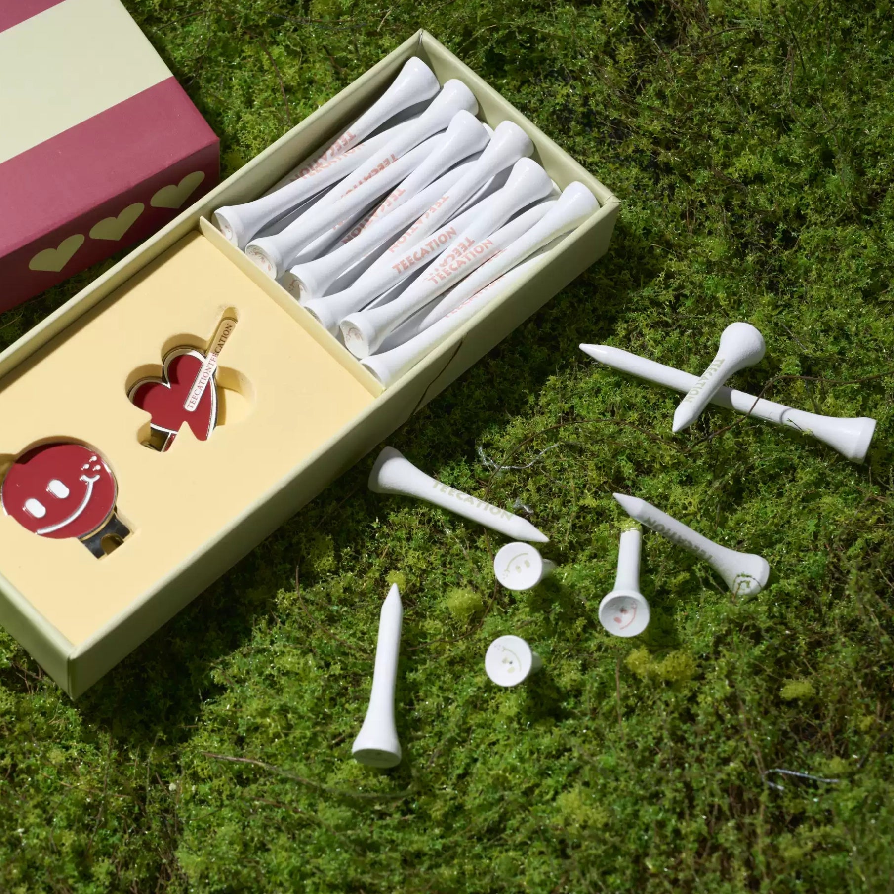 A set of white golf tees and clubs arranged in and around an open decorative gift box on green moss outdoors. The box contains branded golf tees with red text, a heart-shaped pin, and a small round accessory.