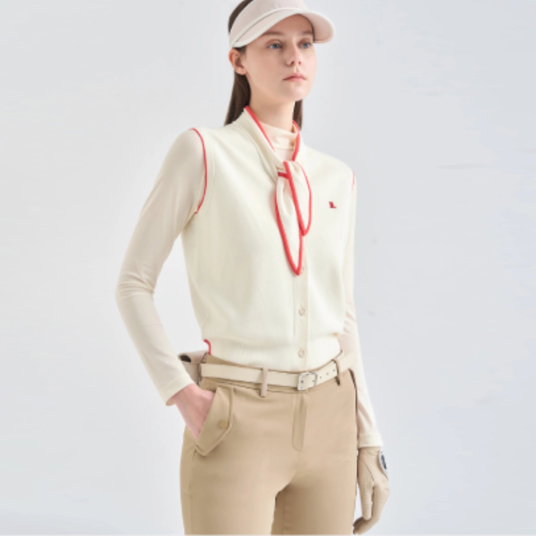 A woman modeling a light cream long-sleeve button-front top with red piping and a matching red neck tie, paired with beige pants and a beige visor against a light gray studio background.