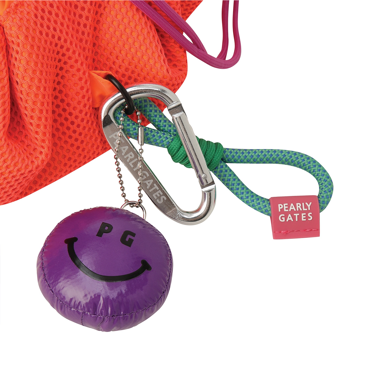 A close-up product shot showing a purple round keychain with a smiley face and the letters PG, attached to a silver carabiner. The carabiner is connected to a green rope and a bright orange mesh drawstring bag.