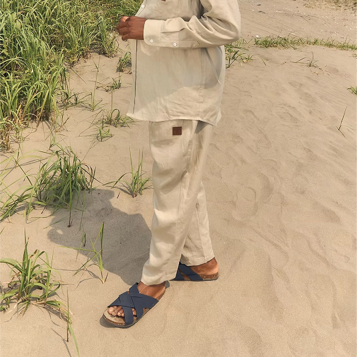 A person standing on sandy ground wearing a light beige linen shirt and matching pants, with dark blue slide sandals. The photo is taken outdoors with grass and sand visible.