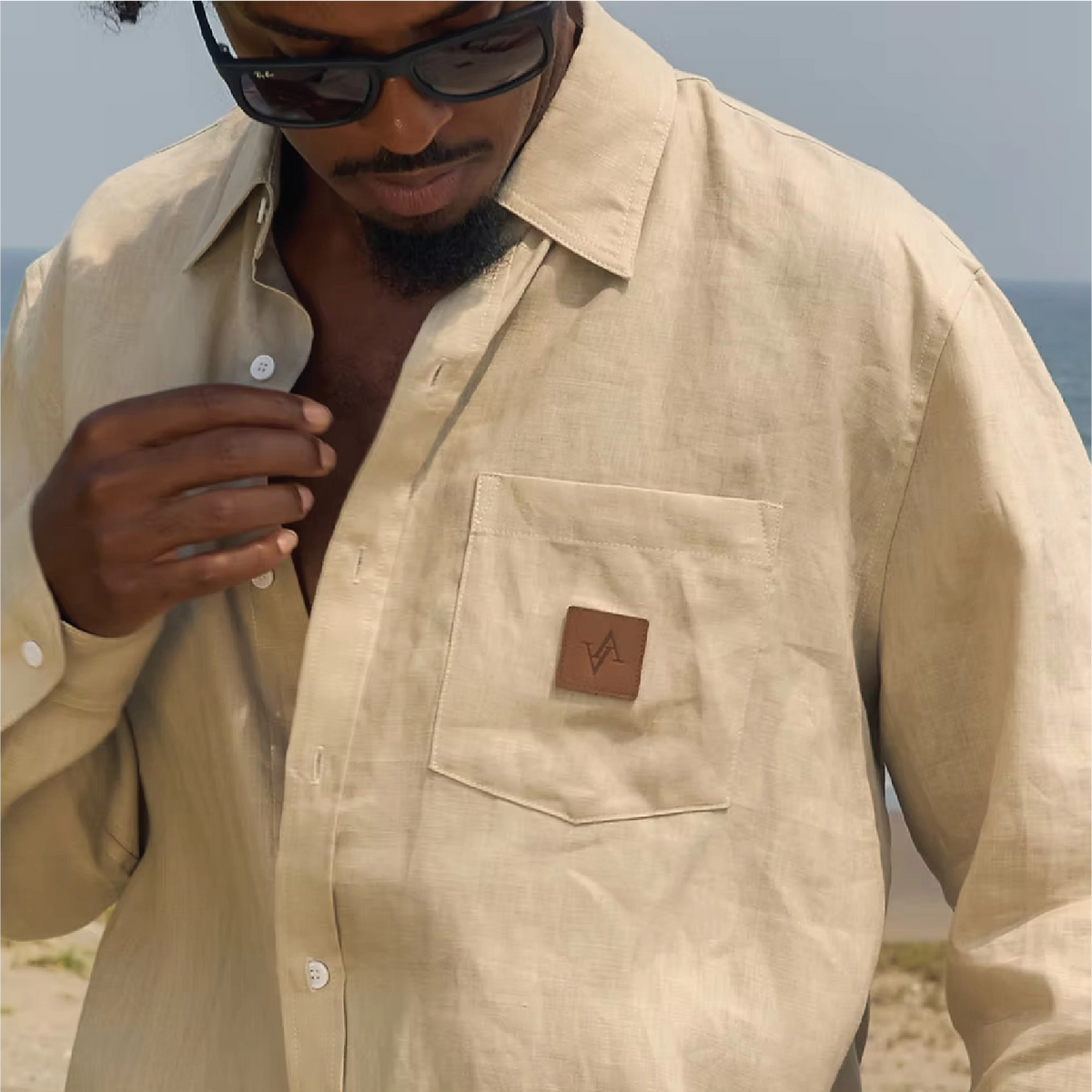 A man wearing a light beige linen button-up shirt with a chest pocket featuring a brown leather brand patch, photographed outdoors near the beach.