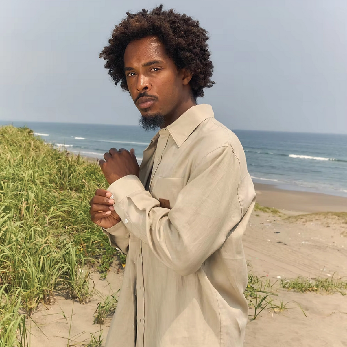 A man with curly dark hair stands on a sandy beach dune wearing a light beige long-sleeve button-up shirt, with the ocean and grass in the background.
