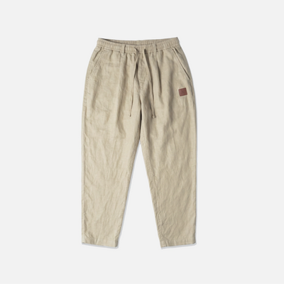 A pair of beige drawstring trousers with a small brown leather patch on the left hip, laid flat against a light gray background.