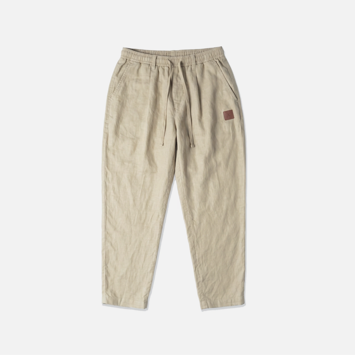 A pair of beige drawstring trousers with a small brown leather patch on the left hip, laid flat against a light gray background.