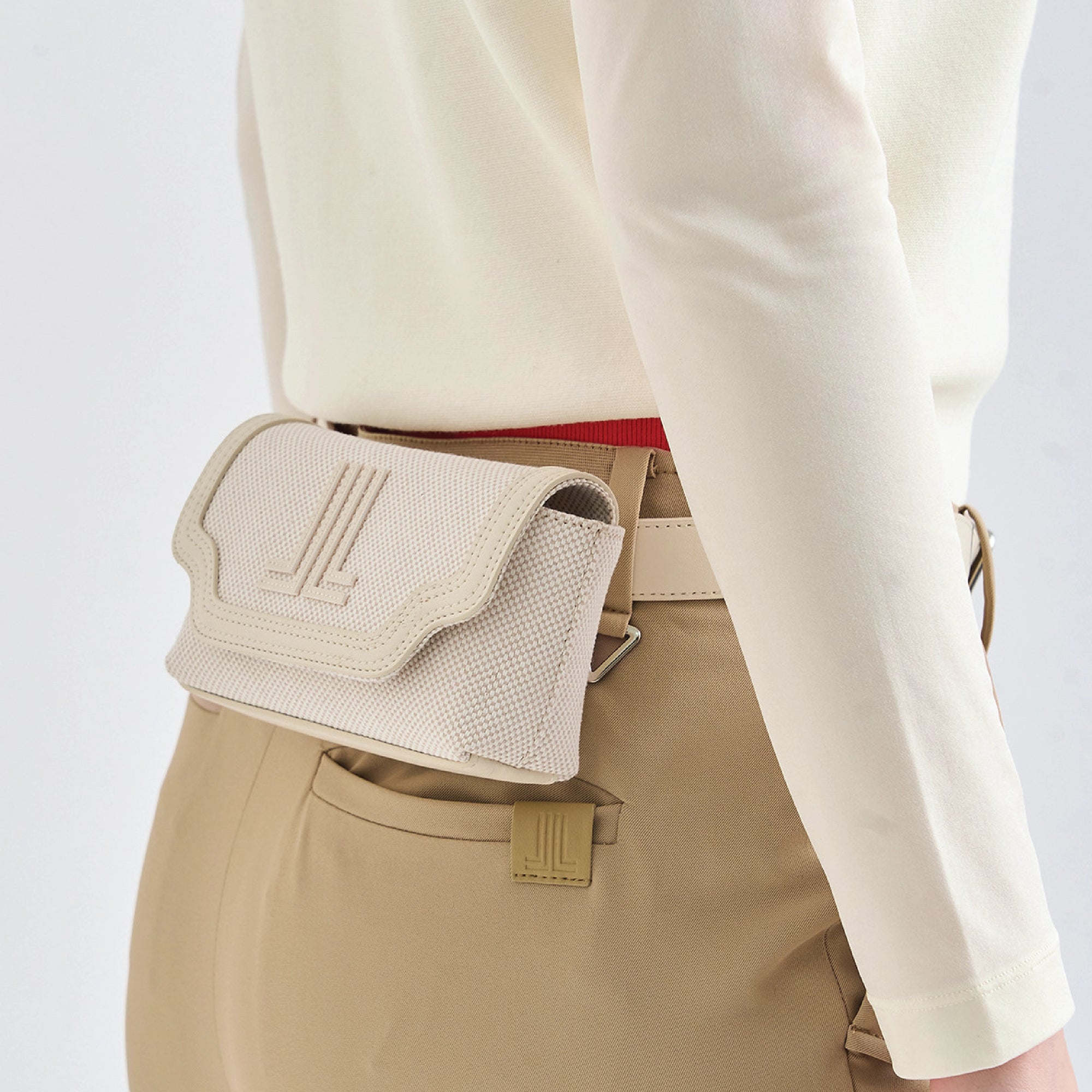 A close-up of a person wearing a beige woven belt bag pouch with an embossed logo, styled with khaki trousers and a cream long-sleeve top.