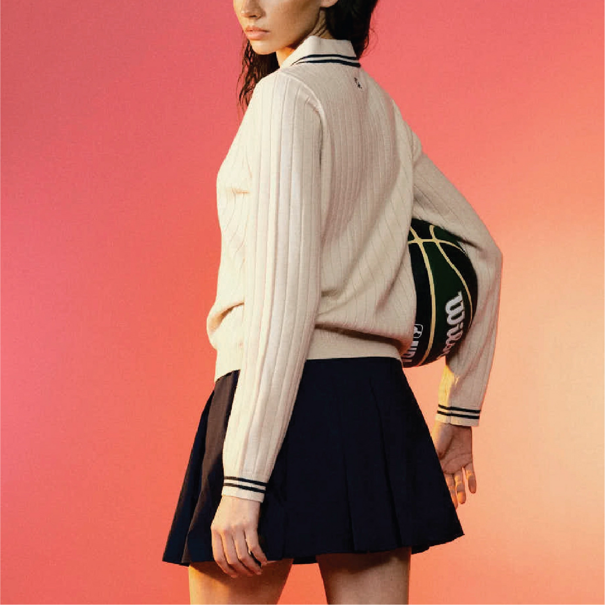 A model shown from the side wears a cream ribbed knit mock-neck sweater with black and white striped cuffs and collar, paired with a dark pleated mini skirt. She is holding a black basketball with yellow lines against a vibrant pink-to-orange gradient background.