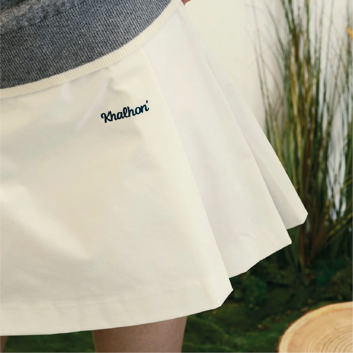 Close-up of a pleated cream-colored skirt with a gray ribbed waistband and the brand name 'Khalhon' embroidered in black script on the side, shown on a model with a blurred green background.