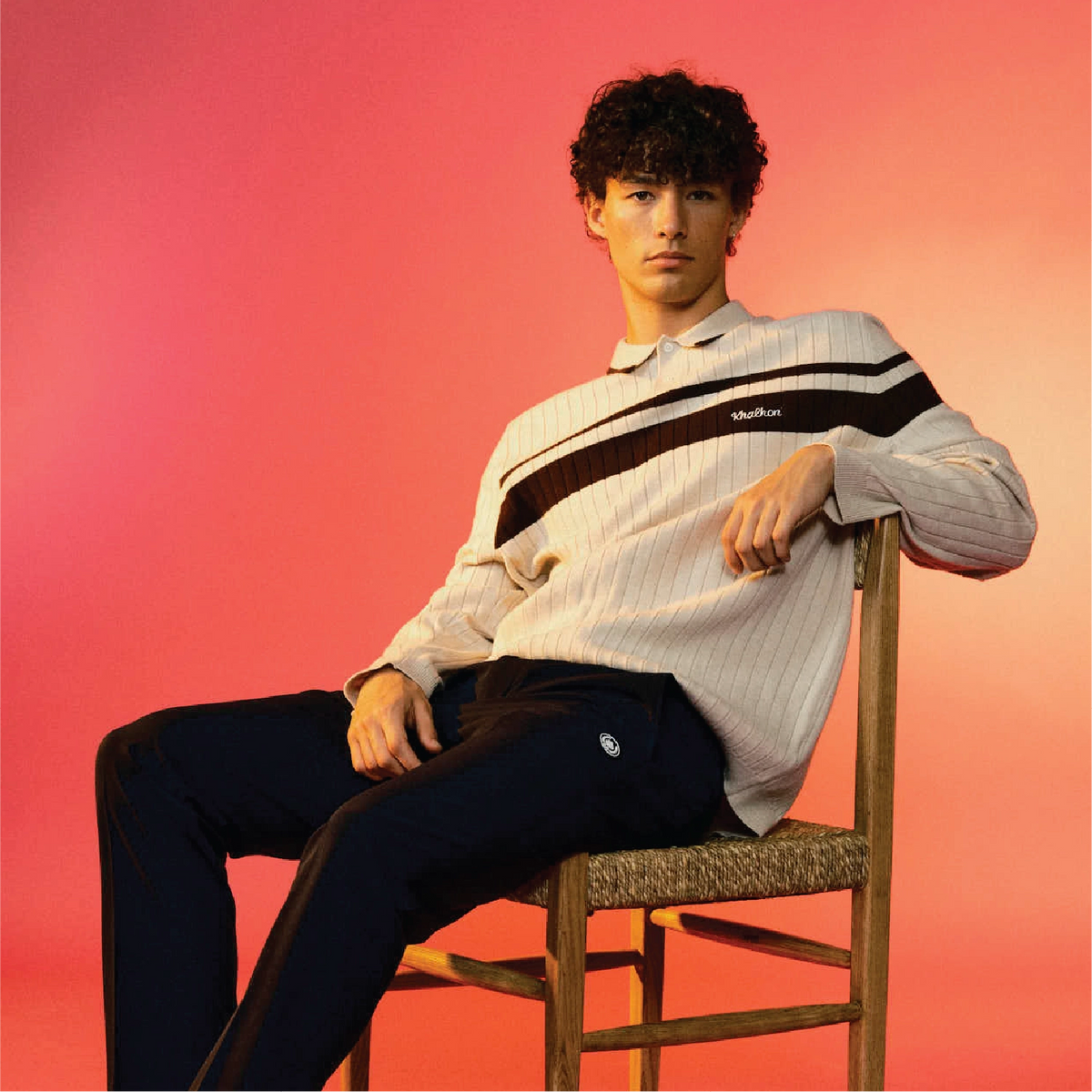 A young man sits on a wooden chair against a vibrant pink and orange gradient background, wearing a ribbed knit polo sweater with black and brown stripes and dark navy pants. The brand name 'Khalhon' is visible on the chest of the sweater.