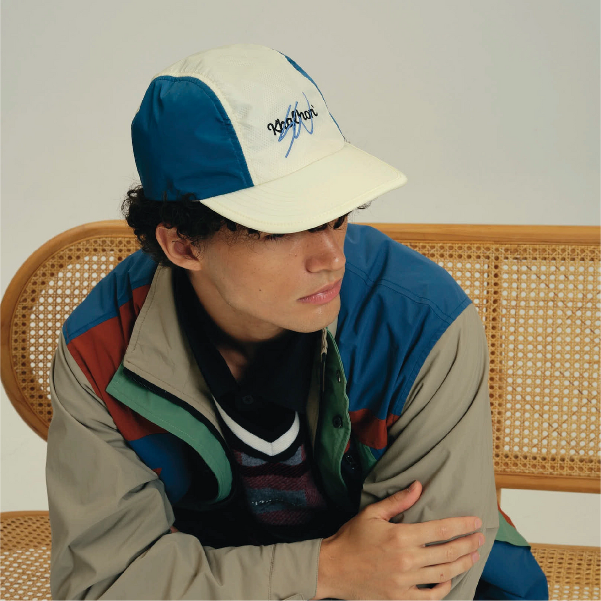 A model wearing a blue and off-white Packeray baseball cap and a multi-colored colorblock jacket, sitting on a woven cane chair against a plain light gray background.