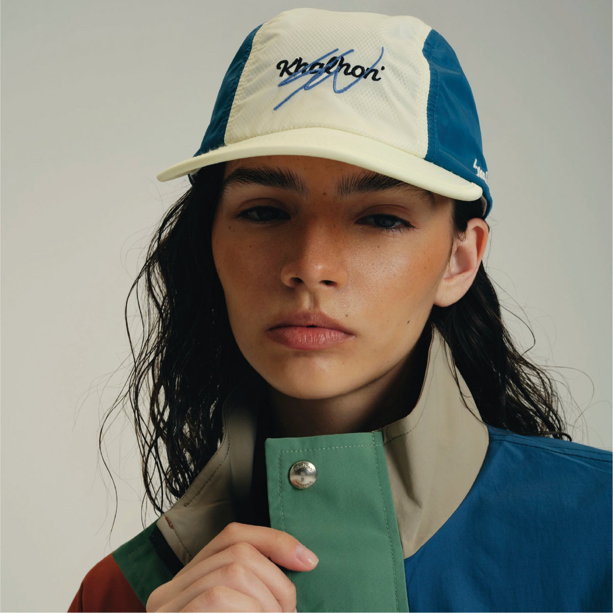 A close-up portrait of a model wearing a two-tone trucker cap with a cream front and blue side panels, featuring the brand name 'Kith' embroidered on the front. The model is also wearing a color-blocked jacket with green, blue, and beige panels.