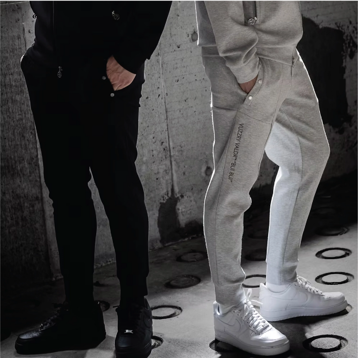 Two models wearing slim-fit jogger sweatpants, one in black and one in light gray, standing against a textured concrete wall. The light gray pants feature a zippered pocket on the side.