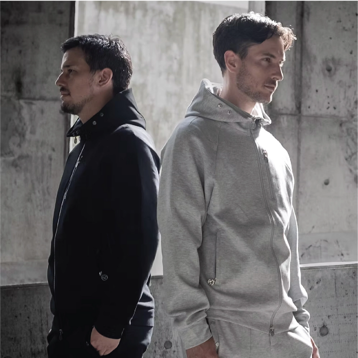 Two men standing back-to-back in a concrete industrial setting, one wearing a black jacket and the other wearing a light gray zip-up hoodie and matching pants.