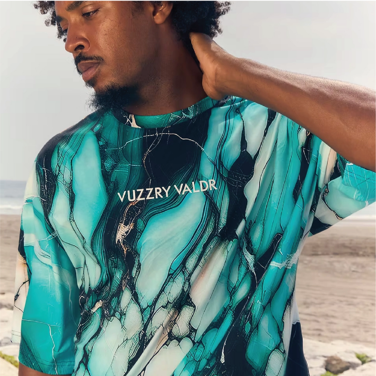 A man wearing a short-sleeve t-shirt with a teal, green, and black marble pattern and the text 'YUZZRY VALDR' printed on the chest, set against a beach background.