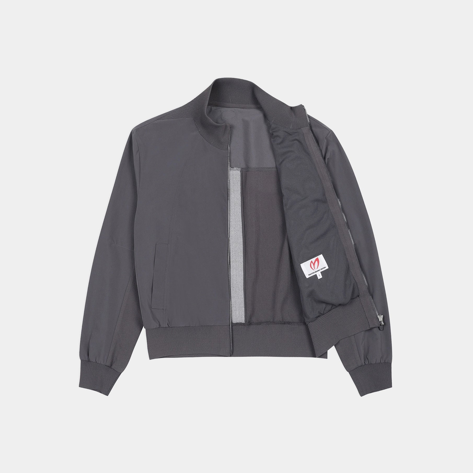 A dark gray zip-up jacket with a high collar, laid flat against a light gray background. The jacket features a vertical lighter gray stripe on the front, a zippered pocket on the left side, and a small white brand patch on the right pocket.