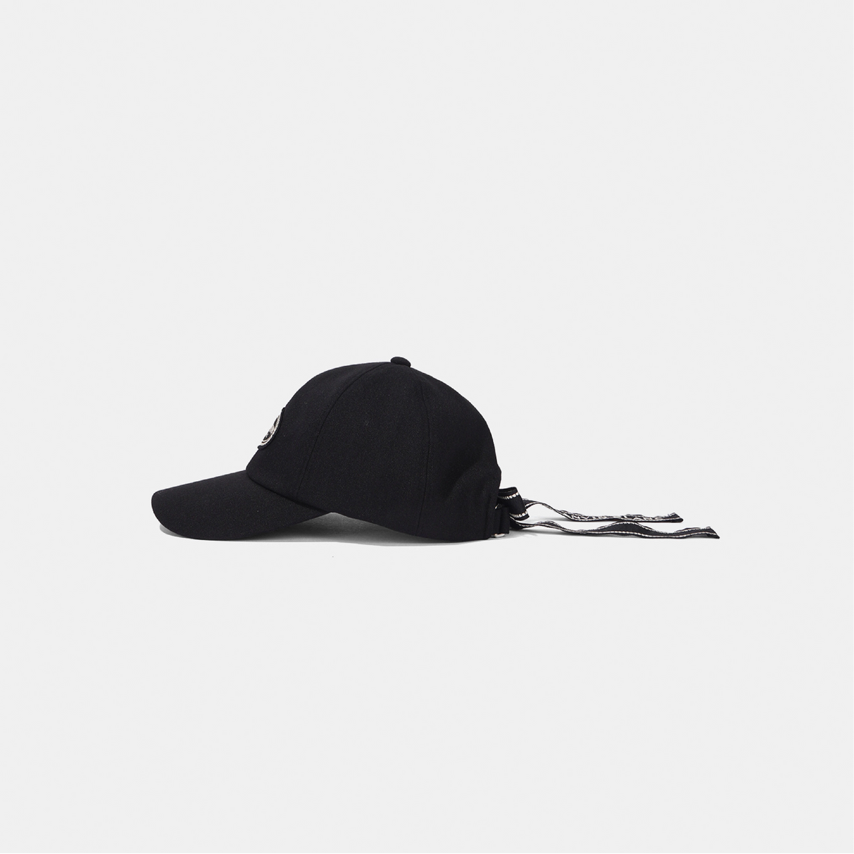 A black baseball cap with a small white embroidered logo on the side, shown in profile against a light gray background. The back of the cap has a drawstring tie with a black and white patterned fabric.
