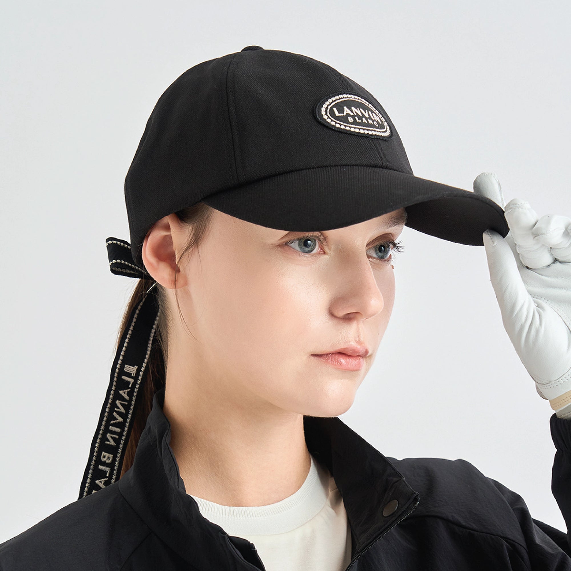 A woman wearing a black baseball cap with a curved brim and a circular embroidered patch on the front. The patch reads 'LANVIN' with 'BLANC' below it.