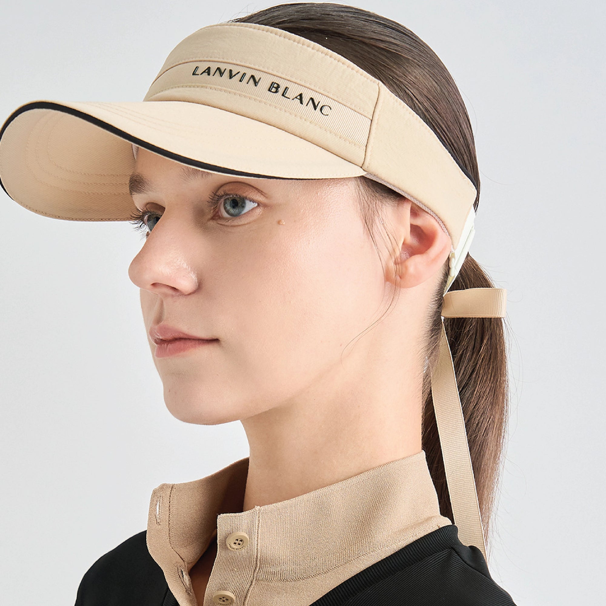 A woman in profile wearing a beige visor with black trim and the brand name 'LANVIN BLANC' printed on it. The visor has a chin strap, and she is wearing a matching beige collared top against a light gray studio background.