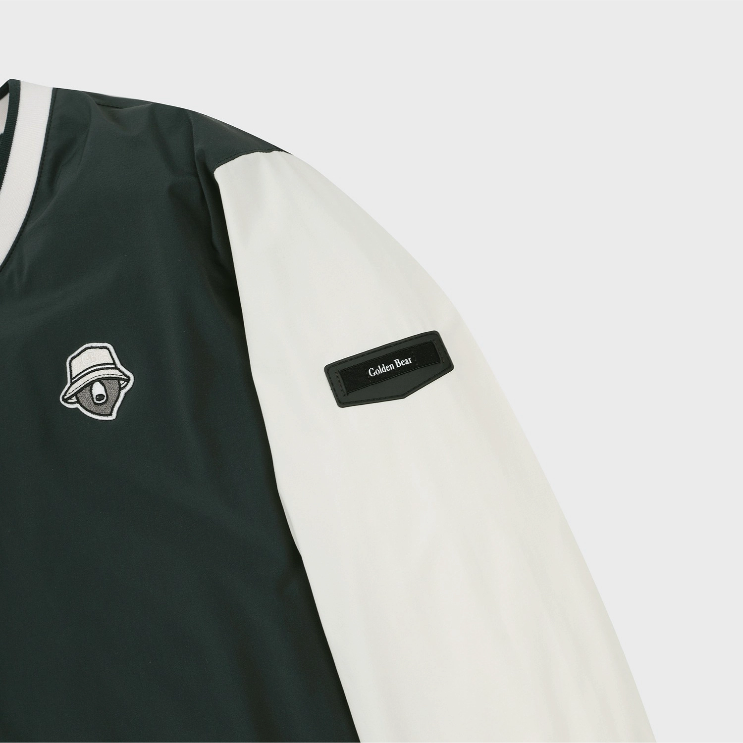 Close-up of a color-blocked jacket sleeve and chest area, featuring a dark forest green body, an off-white sleeve, a black 'Galen Bear' brand patch, and a small embroidered bear logo on the chest.