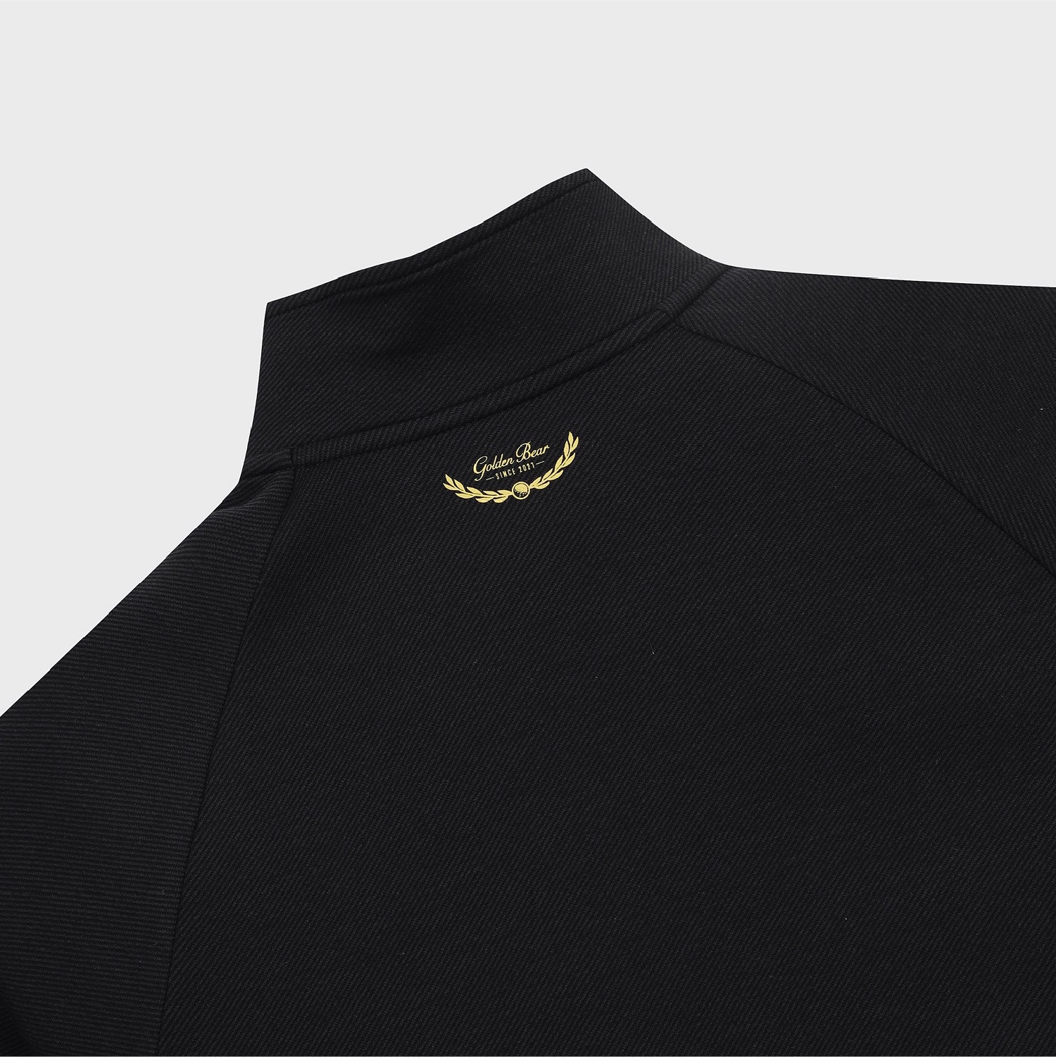 Close-up of a black textured pullover top with a mock neck collar, featuring a small gold embroidered laurel wreath and 'Golden Bear' text on the upper chest, set against a light gray background.