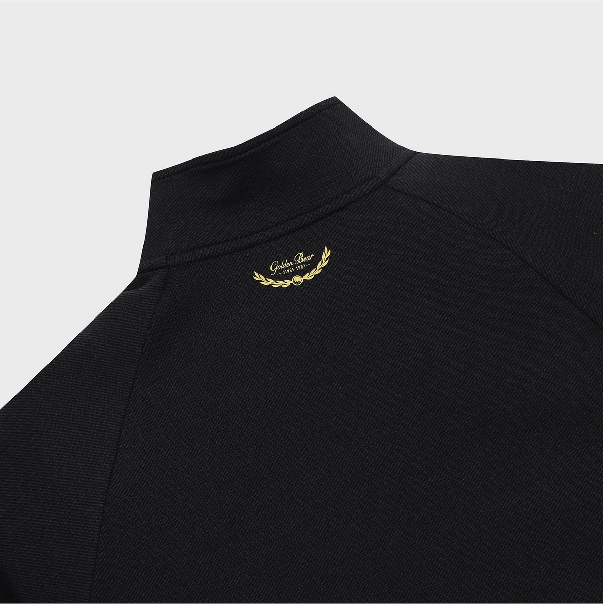 Close-up of a black textured pullover top with a mock neck collar, featuring a small gold embroidered laurel wreath and 'Golden Bear' text on the upper chest, set against a light gray background.