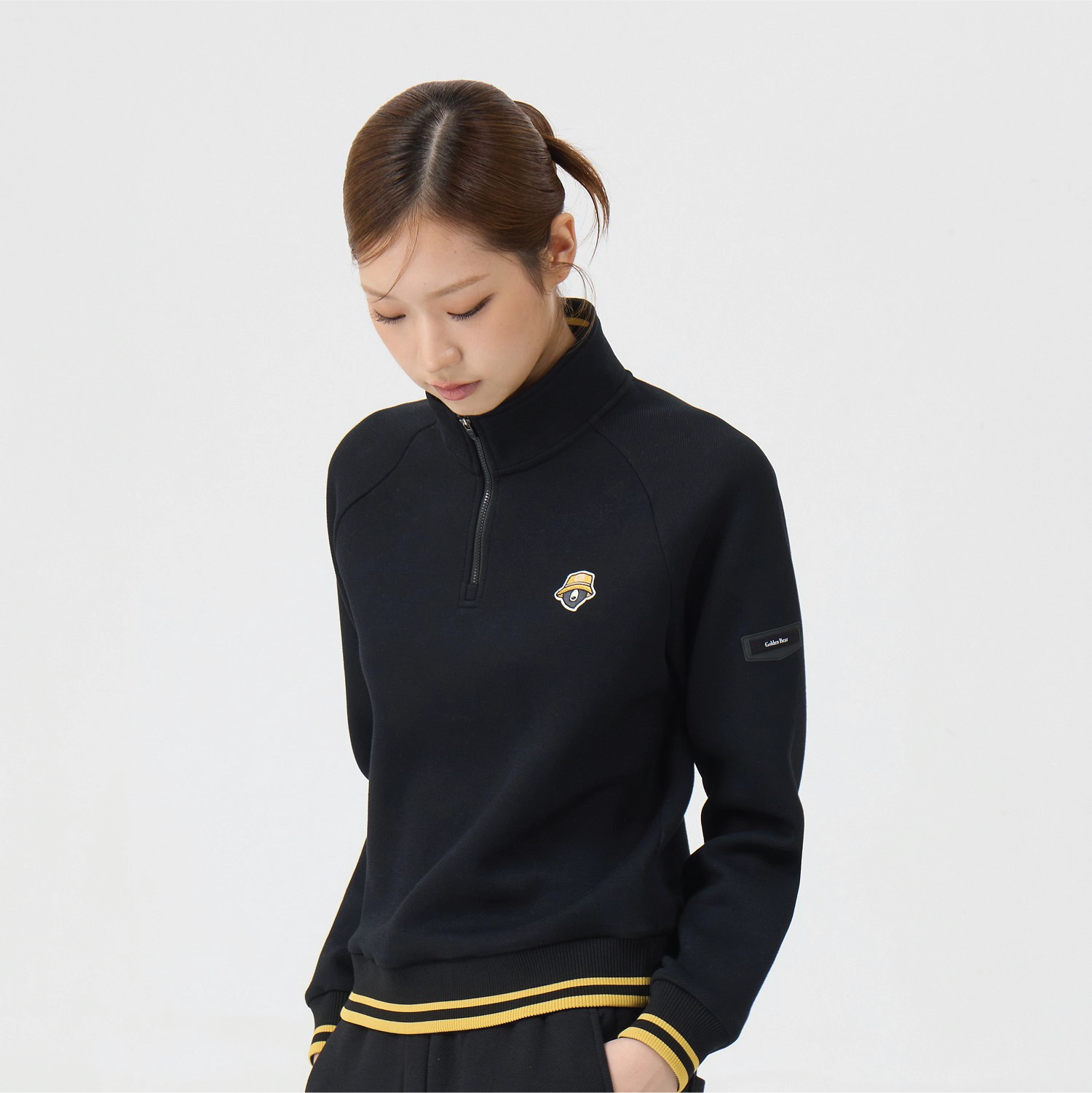 A woman wearing a black long-sleeve quarter-zip pullover with a small chest logo and a sleeve patch, paired with matching black bottoms with yellow stripes, against a light gray studio background.
