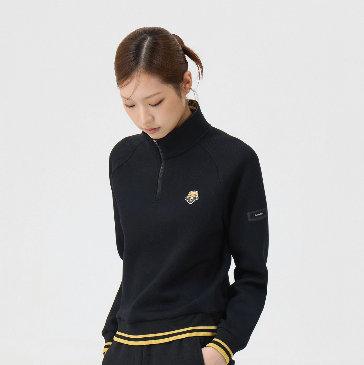 A woman wearing a black long-sleeve quarter-zip pullover with a small chest logo and a sleeve patch, paired with matching black bottoms with yellow stripes, against a light gray studio background.