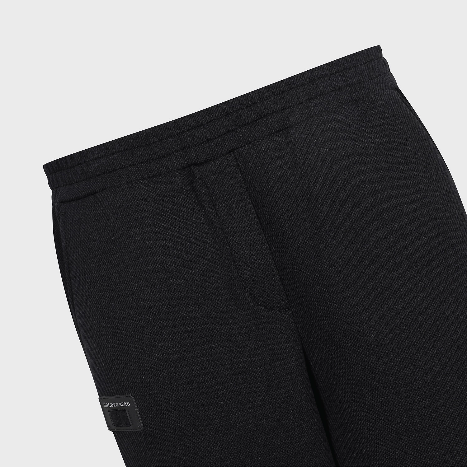 Close-up of black textured sweatpants showing the elastic waistband, side pocket, and a small black brand patch near the hem on a light gray background.