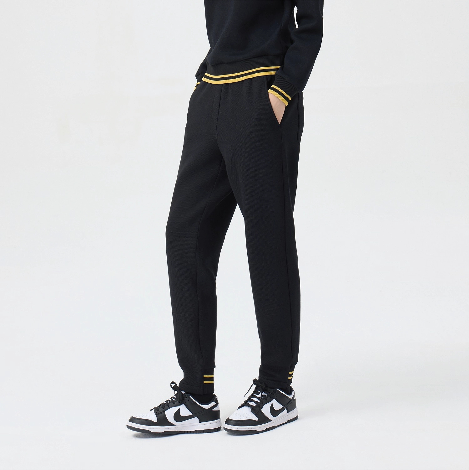 A person wearing black jogger-style pants with yellow stripe accents at the waist and cuffs, paired with a black top and black-and-white sneakers, photographed against a clean light gray studio background.