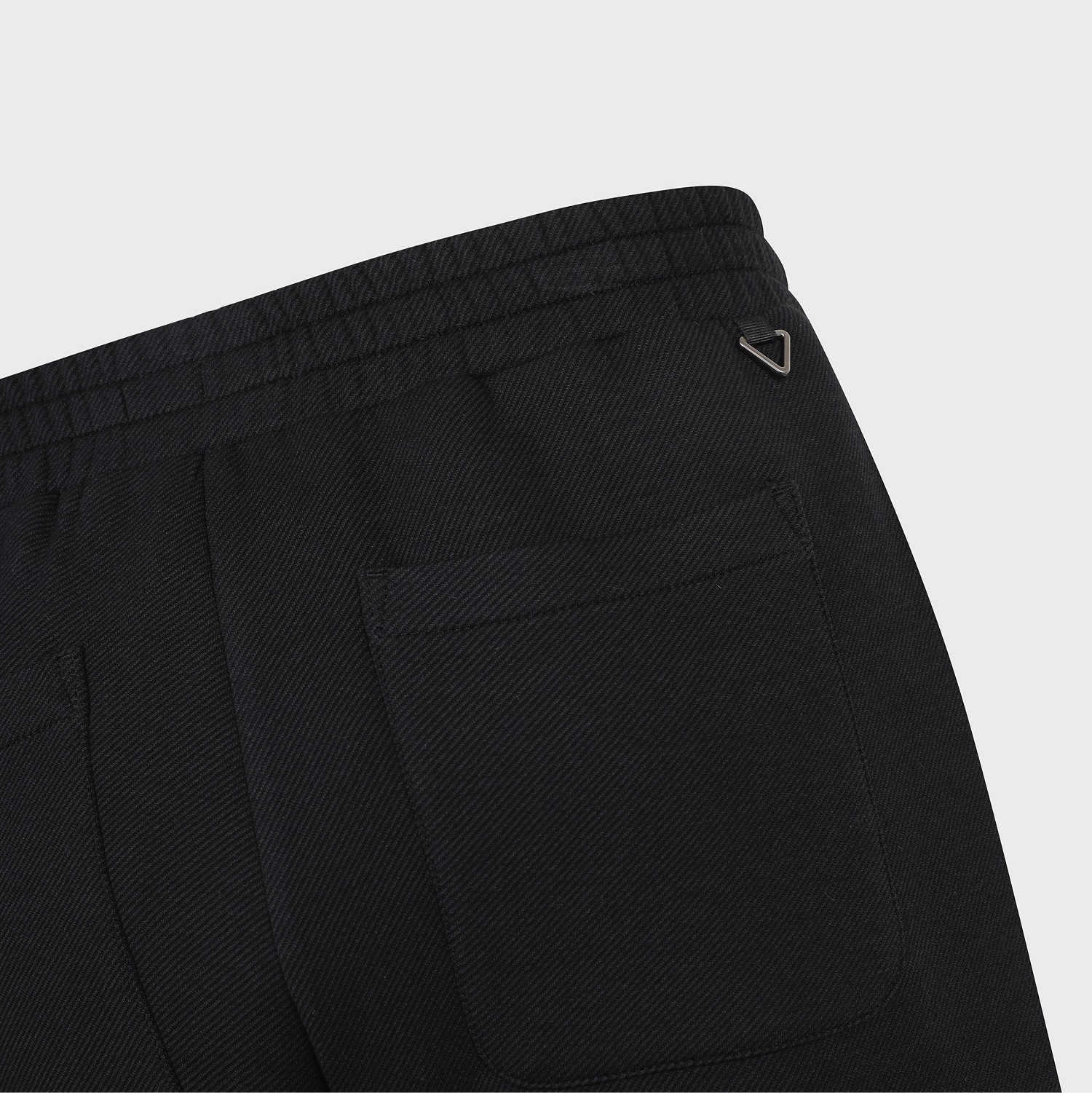 Close-up of the back waistband and pocket area of black textured trousers, showing a small metallic logo tab on the upper right side against a light gray background.