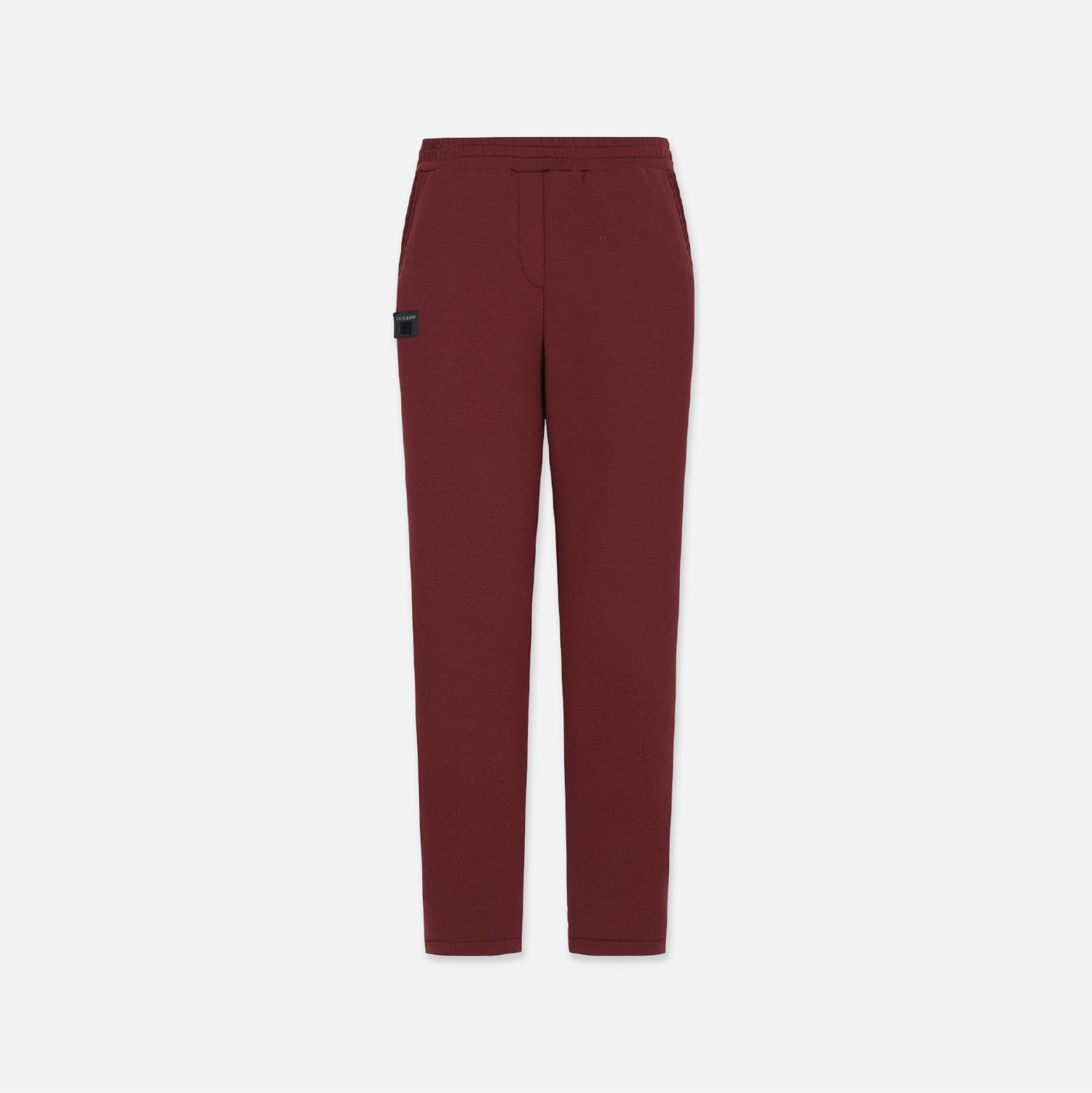 A pair of slim-fit trousers in a deep burgundy red, featuring a small black rectangular patch on the left thigh, displayed against a solid light gray background.