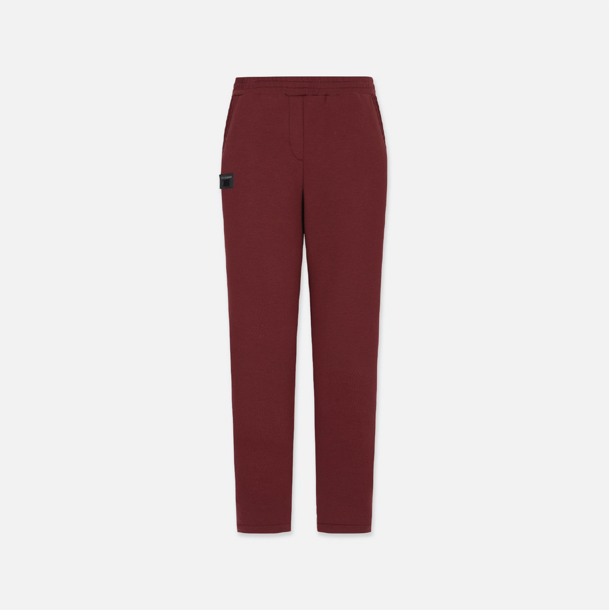 A pair of slim-fit trousers in a deep burgundy red, featuring a small black rectangular patch on the left thigh, displayed against a solid light gray background.