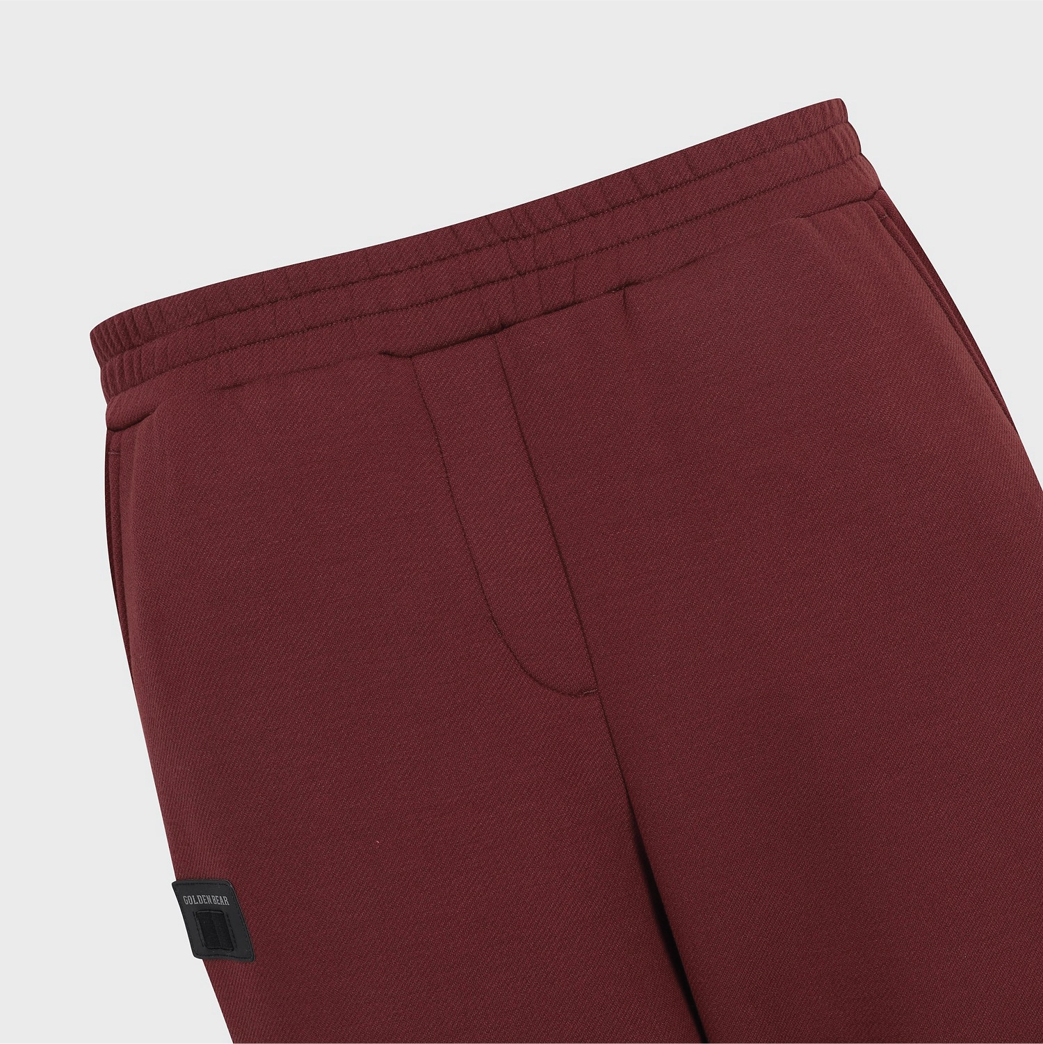 Close-up of burgundy fleece sweatpants showing the elastic waistband, side pocket seam, and a small black brand patch on the lower left leg.
