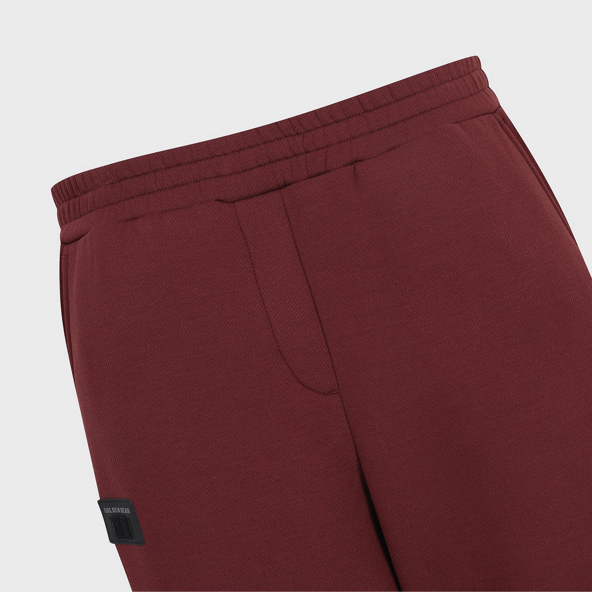 Close-up of burgundy fleece sweatpants showing the elastic waistband, side pocket seam, and a small black brand patch on the lower left leg.
