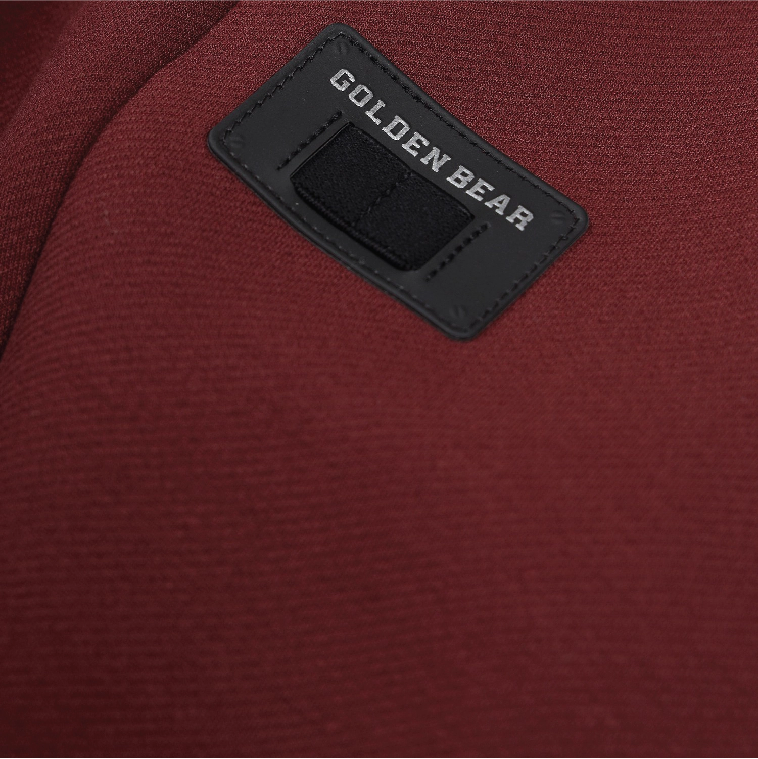 Close-up of a dark red textured fabric with a black rectangular patch stitched onto the surface. The patch has white text reading GOLDEN BEAR and a black hook-and-loop closure.