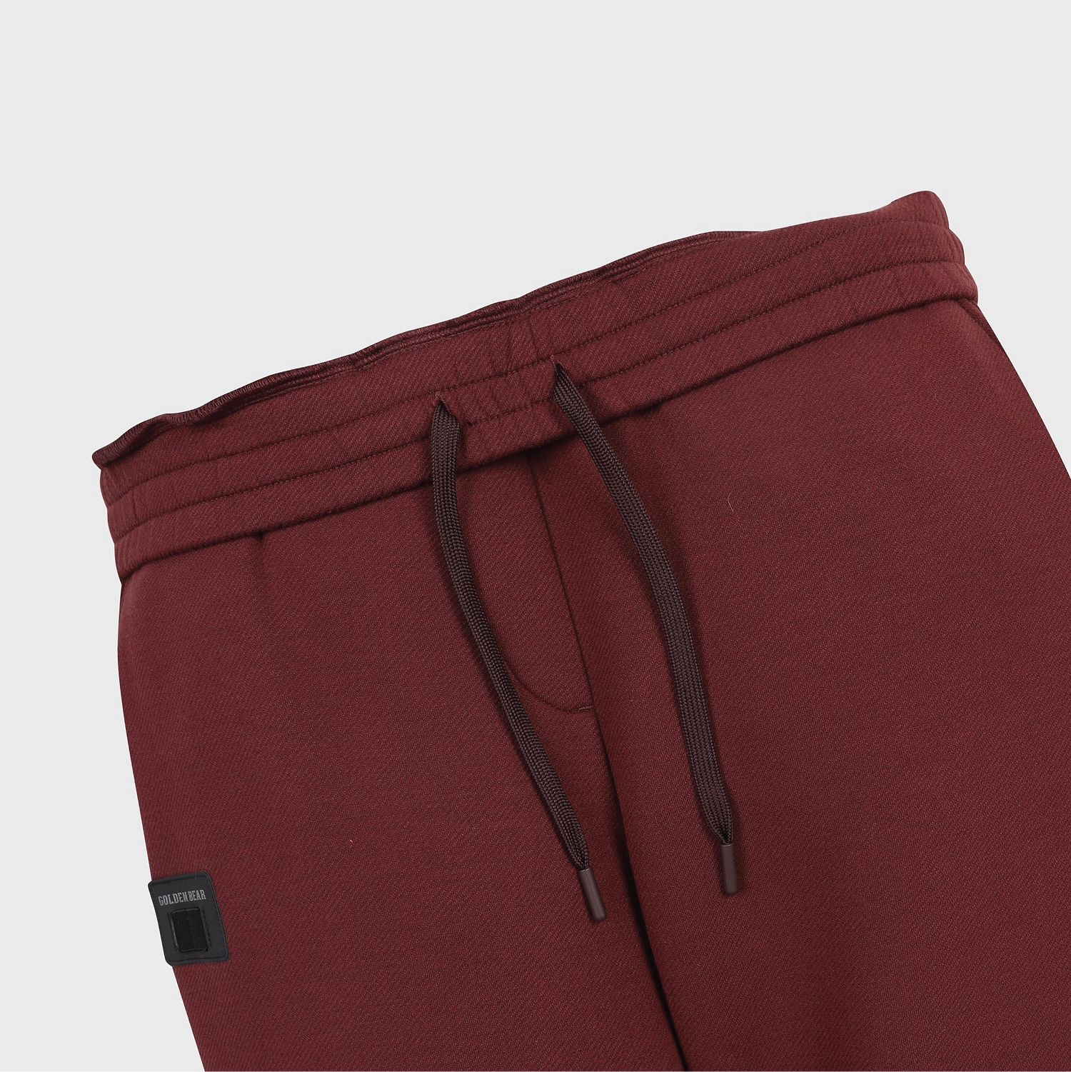 Close-up of burgundy sweatpants showing the elastic waistband, dark brown drawstrings, and a small black brand patch on the left side.