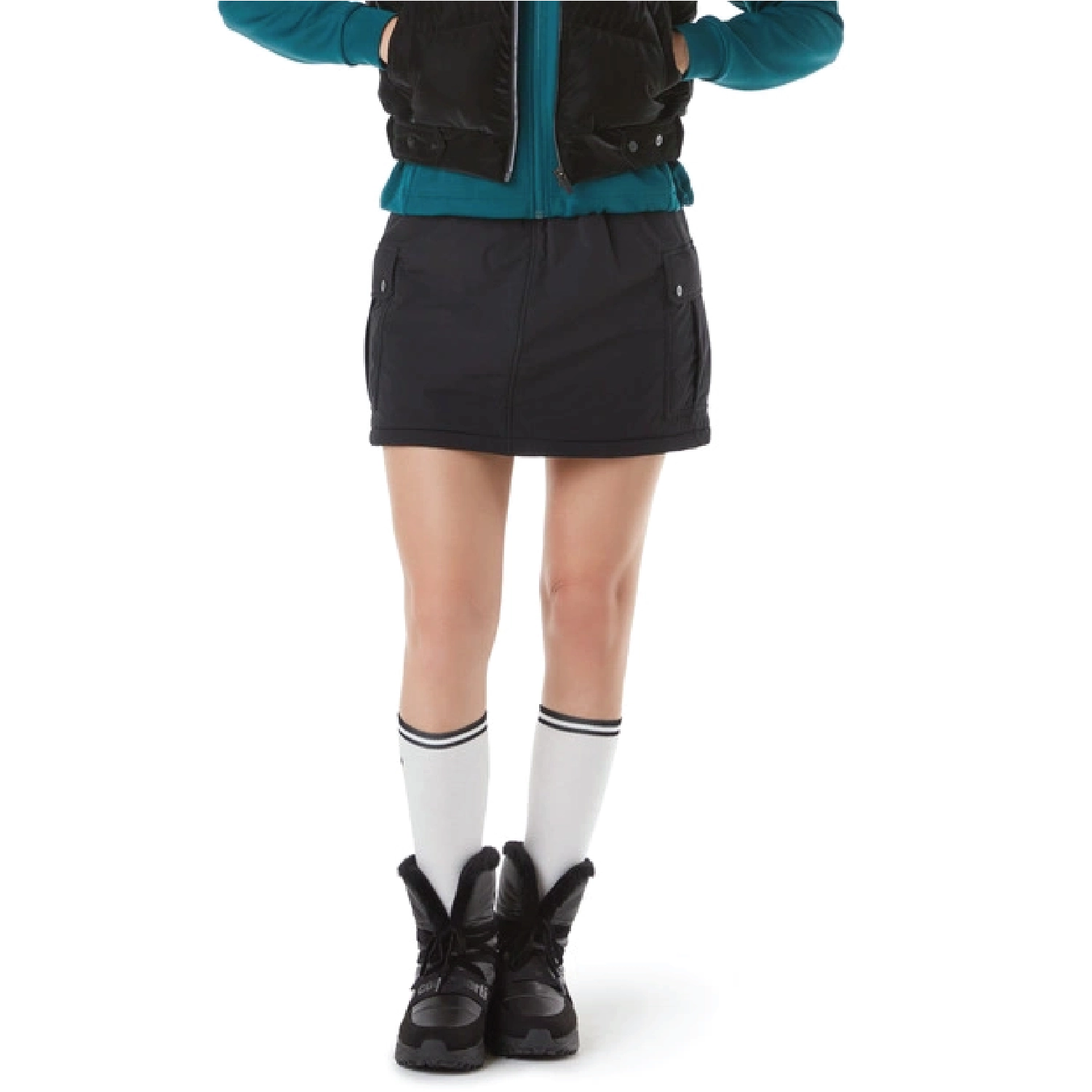 A model is shown from the chest down wearing a black utility-style mini skirt with side pockets, a teal zip-up jacket, a black quilted vest, white knee-high socks with black stripes, and black lace-up boots, against a plain white background.