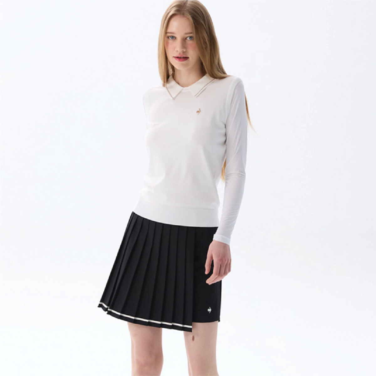 A young woman modeling a white long-sleeve polo knit top and a black pleated tennis skirt with white trim, set against a plain white studio background.