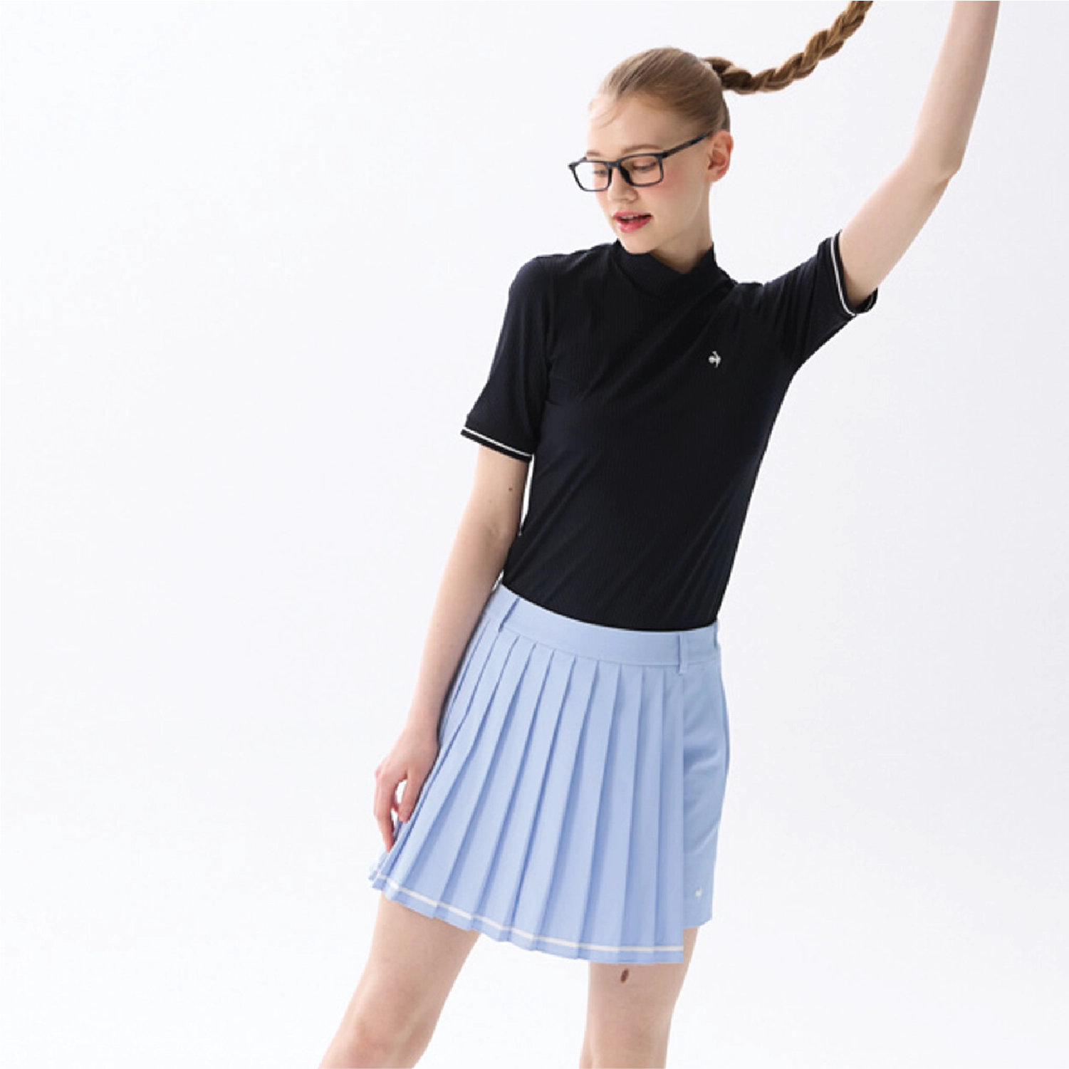 A woman modeling a black short-sleeve mock-neck athletic top with white piping and a light blue pleated skirt, posed against a clean white studio background.