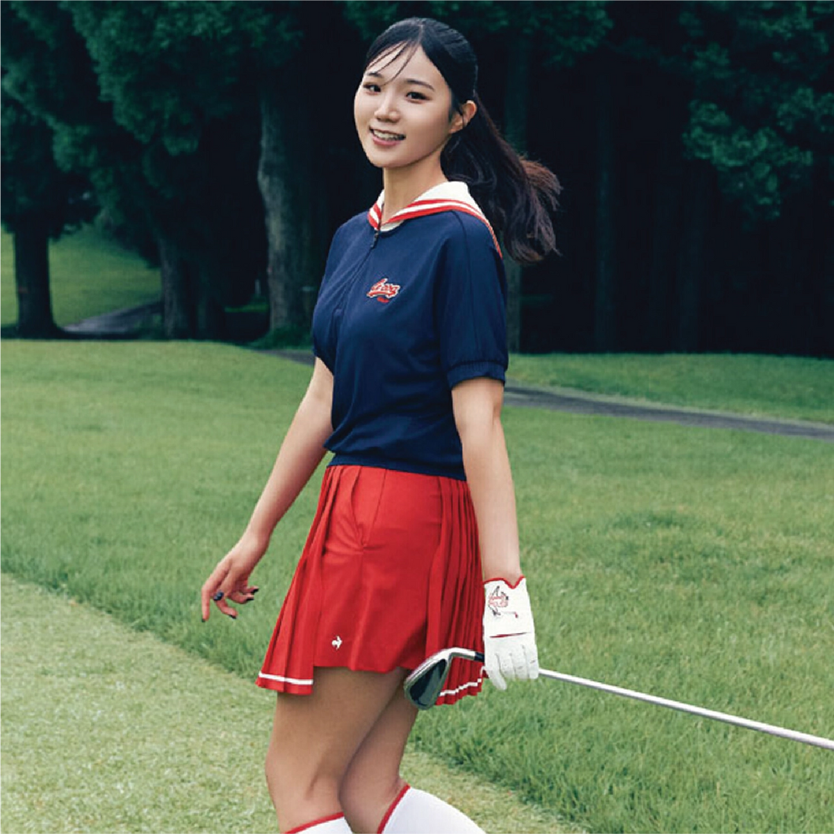 A woman stands on a grassy golf course wearing a navy blue short-sleeve polo shirt with red and white trim, a matching pleated red skirt, white socks, and a white golf glove, holding a golf club.