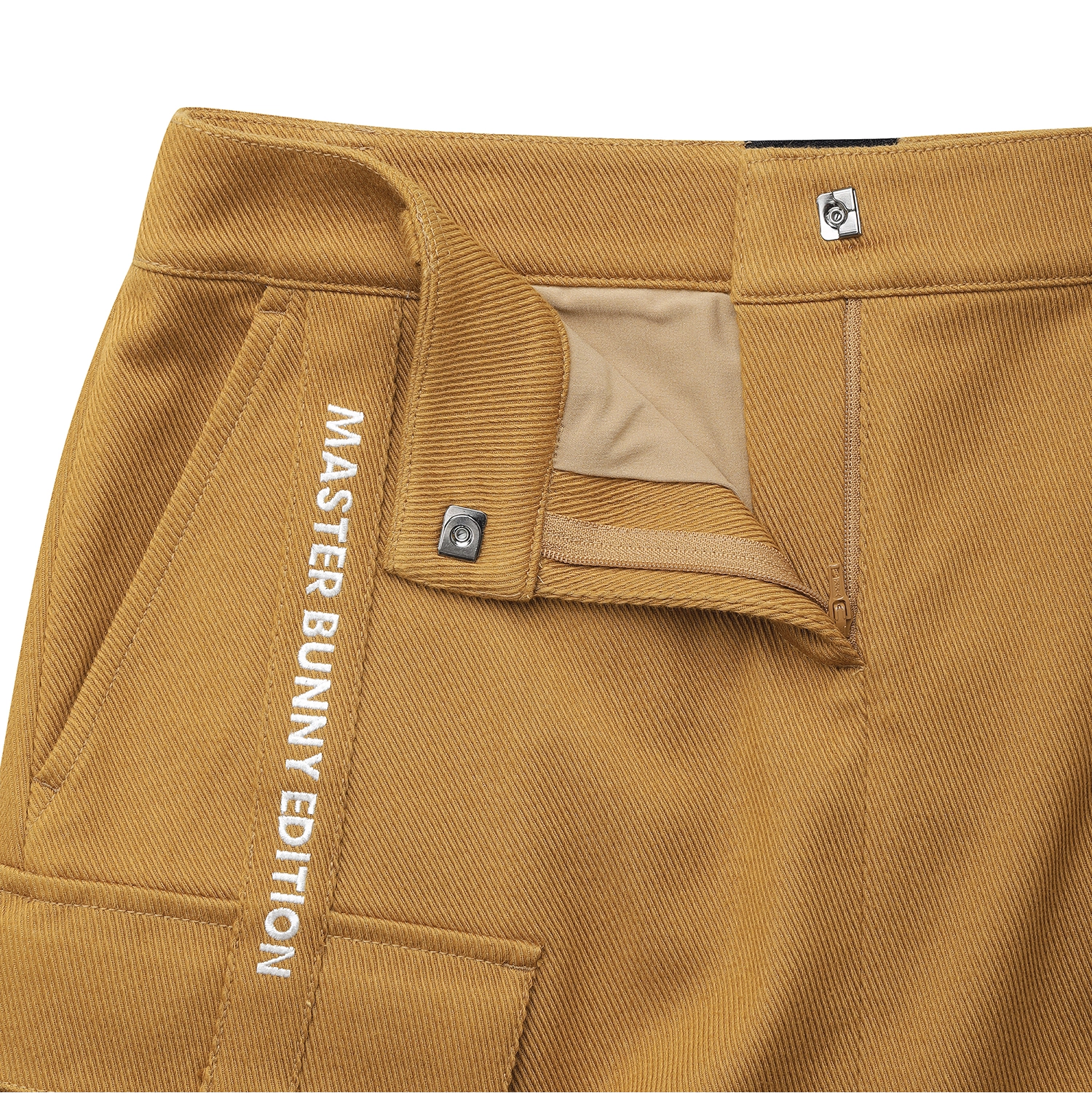 Close-up of mustard yellow trousers showing the waistband, belt loop, pocket edge, and white vertical text reading 'MASTER BUNNY EDITION' on the side.