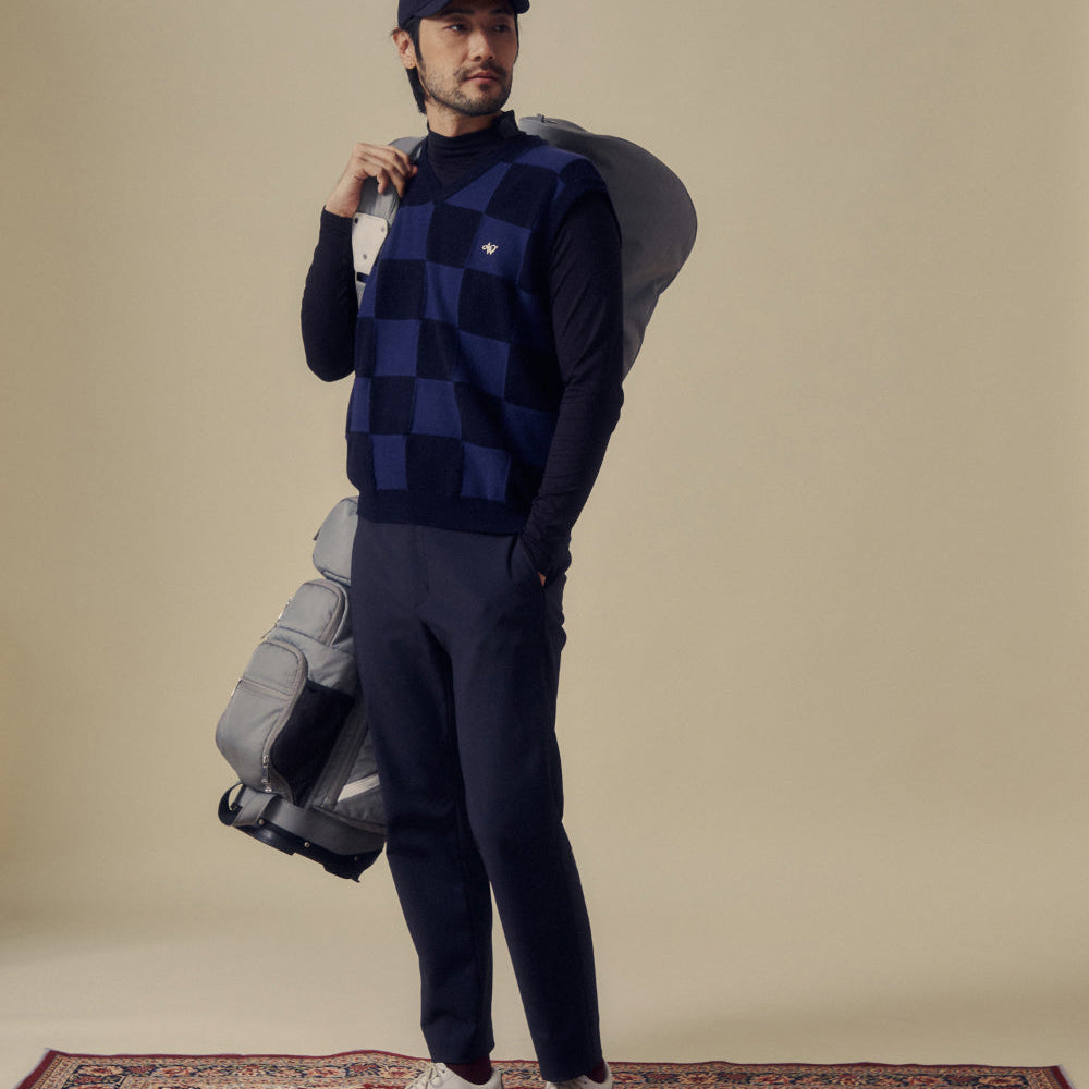 A man modeling a navy blue and dark blue checkered sweater vest over a dark long-sleeve shirt, paired with dark trousers and a dark cap, while carrying a gray golf bag against a plain beige studio background.