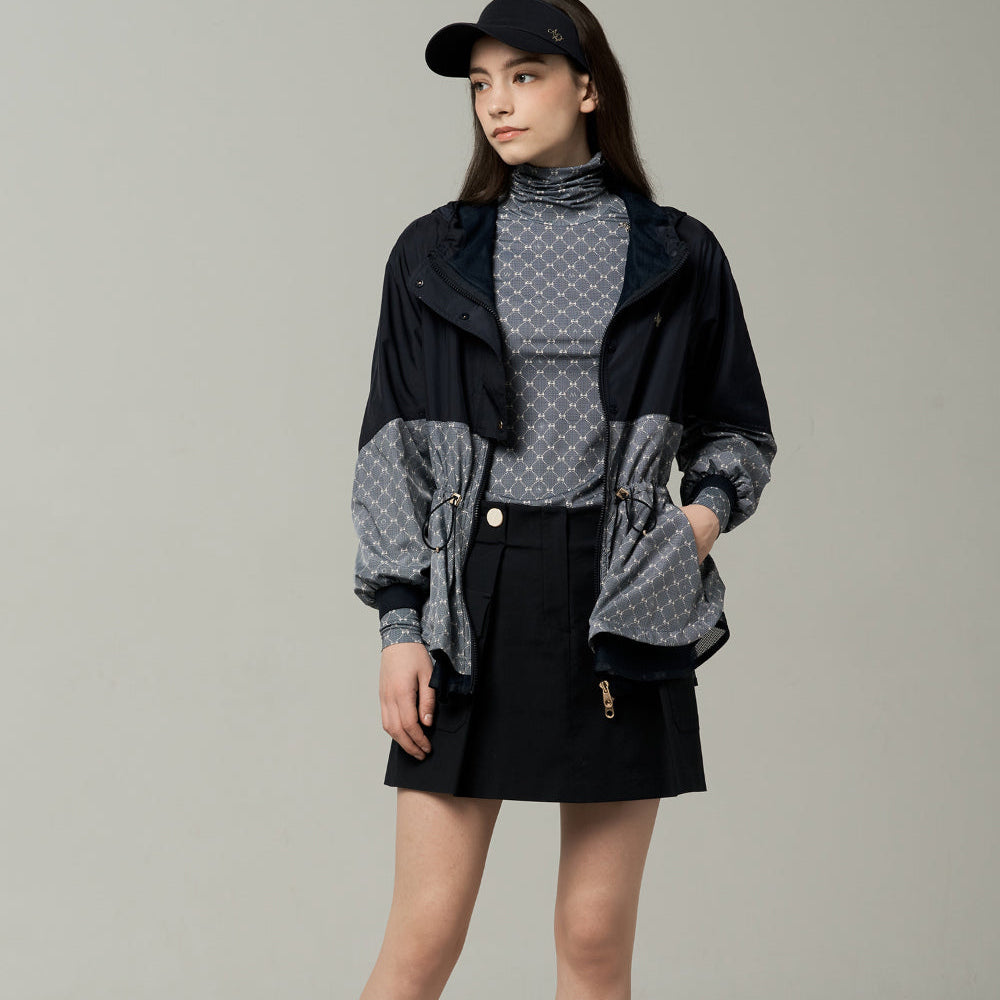 A model stands against a neutral light gray studio background wearing a black and gray patterned windbreaker jacket over a gray patterned mock-neck top, a black mini skirt, white socks, and black-and-white sneakers.