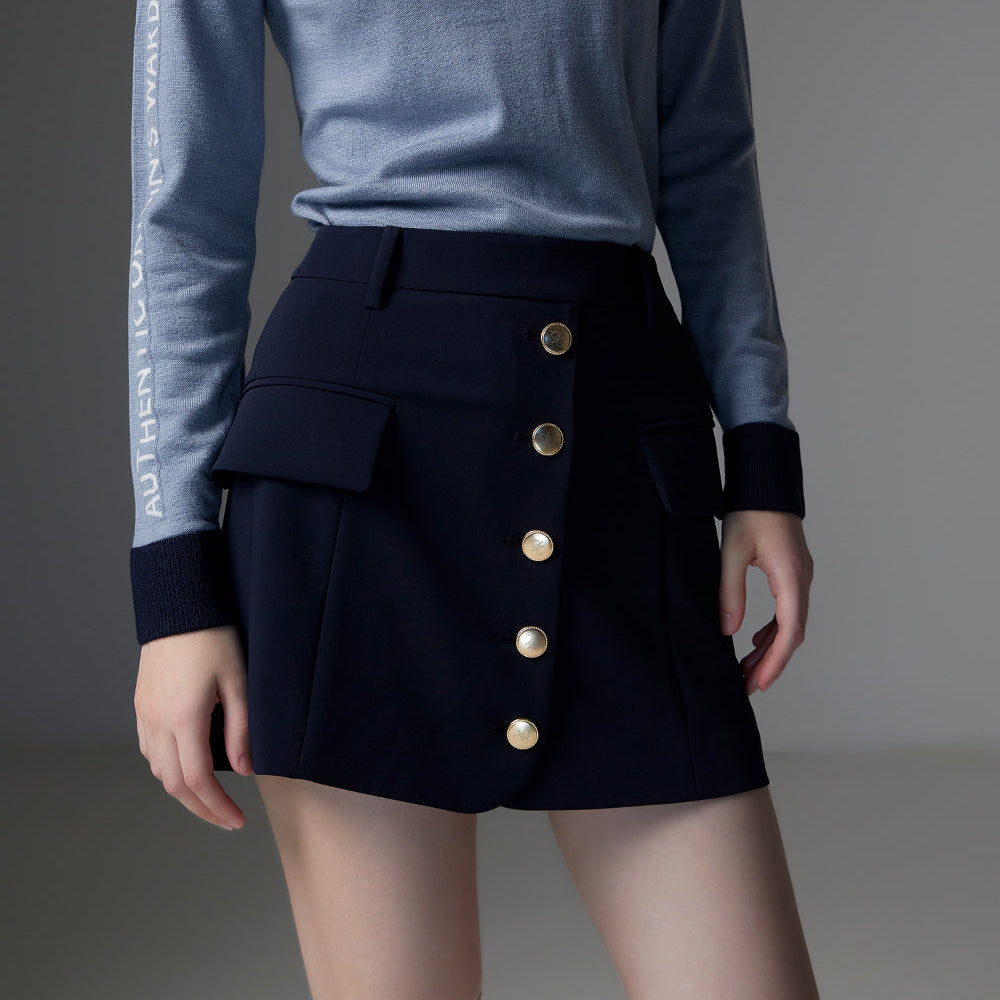 A model wearing a light blue long-sleeve knit top with white text down one sleeve and a dark navy high-waisted mini skirt with gold buttons, paired with white socks.