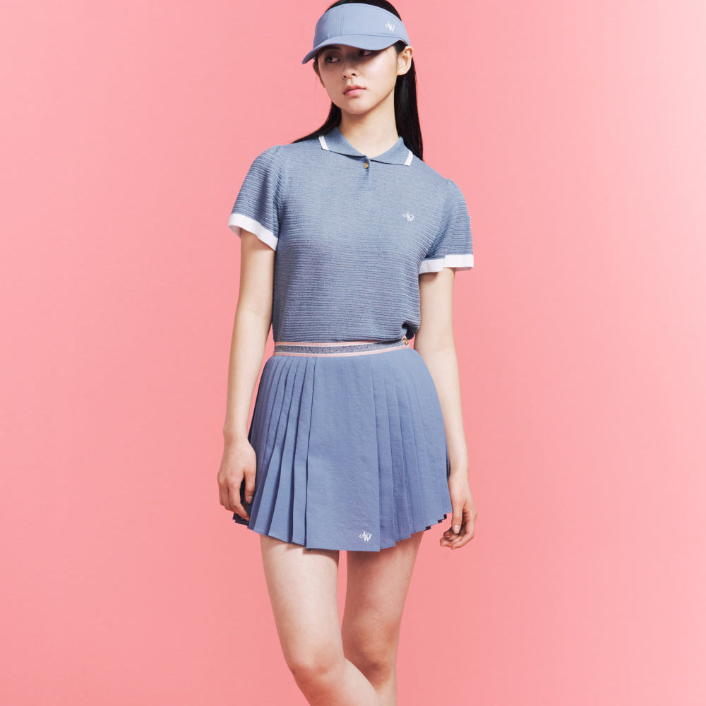 A woman modeling a matching blue knit polo shirt and pleated tennis skirt set with white trim, paired with a light blue visor, against a solid pink background.