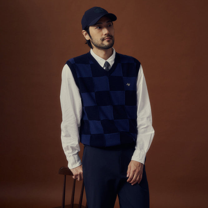 A man wearing a navy blue argyle sweater vest over a white collared shirt and dark trousers, standing against a solid dark brown studio background.