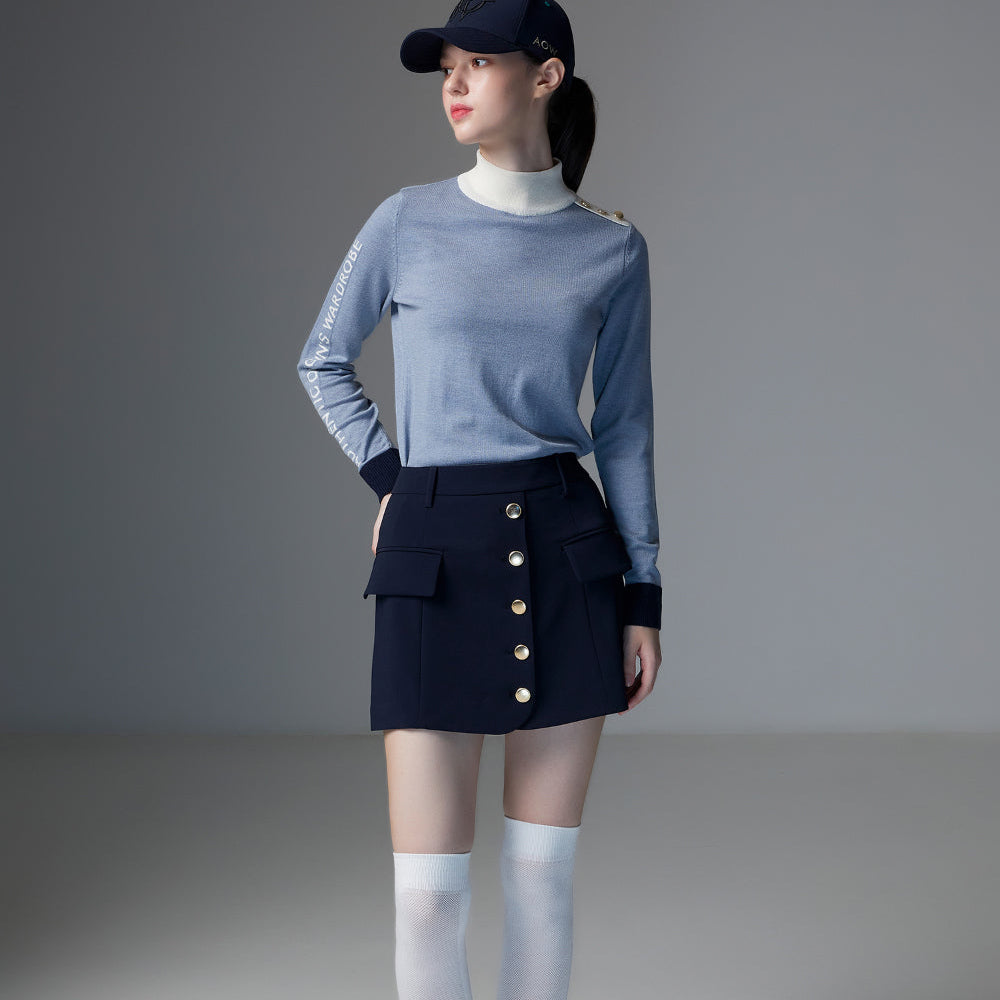 A model stands in a studio wearing a light blue long-sleeve knit top, a dark navy mini skirt with gold buttons, white knee-high socks, and white sneakers, along with a dark navy baseball cap.
