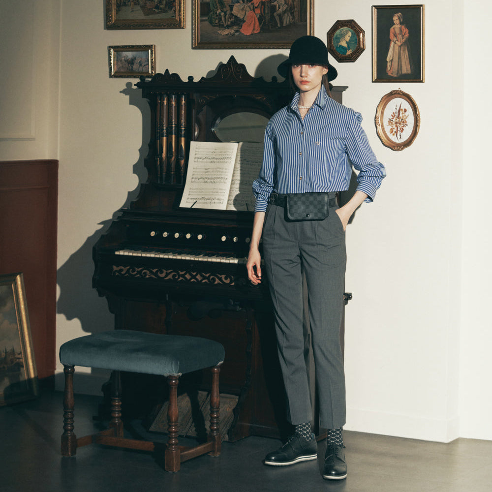 A model stands in a room with a vintage piano and framed wall art, wearing a light blue button-up shirt, dark gray trousers, a black hat, and a black quilted crossbody bag.