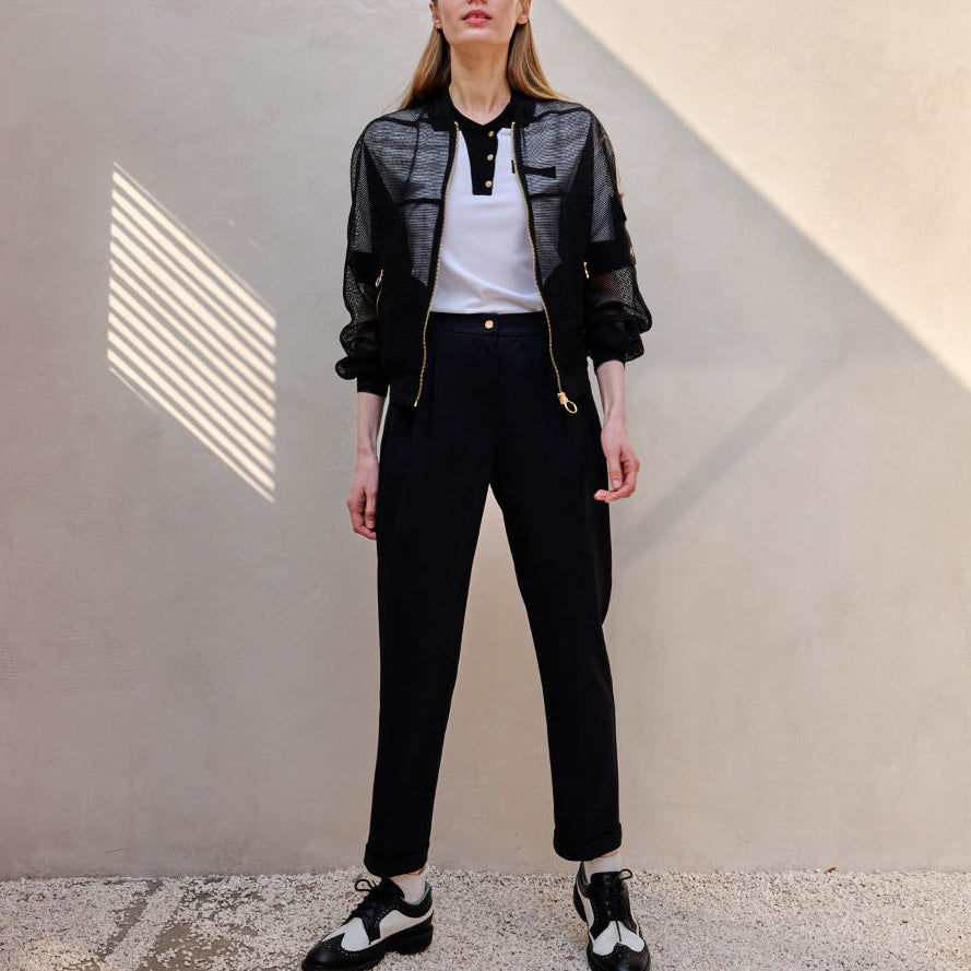 A model stands against a textured beige wall wearing a black sheer mesh zip-up jacket over a white polo shirt, black trousers, and black-and-white shoes.